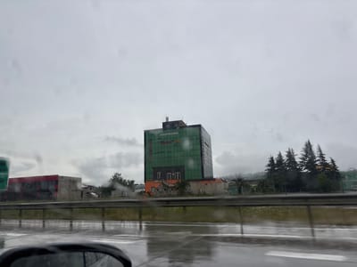 The image, taken from a vehicle on a rainy day in Kveda Simoneti, Georgia, shows a modern, multi-story building with a green glass facade. The building has a prominent sign with text in Georgian script. At its base, the building features an orange section with visible windows and some stone-textured architectural elements. To the left of this building, a smaller, red-accented structure is partially visible. In the background, a line of coniferous trees stands against a cloudy sky. The road in the foreground is wet, reflecting the overcast conditions. A car's side mirror is visible in the lower left corner of the frame.