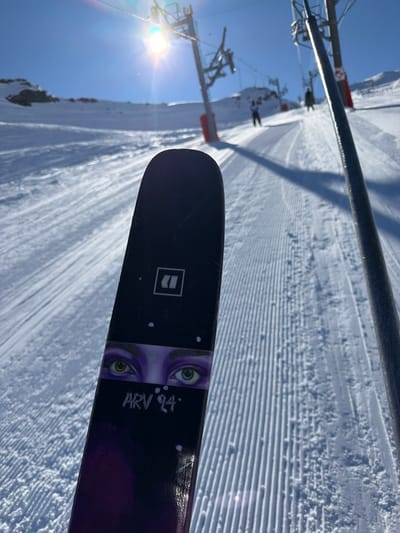 A close-up shot shows the tip of a pair of Armada ARV 94 skis on a ski lift at Saint-Martin-de-Belleville, France. The skis have a black top sheet with a graphic of eyes and the text "ARV 94". The ski lift and a groomed slope covered in snow are visible in the background. The sky is clear and blue. A chairlift tower and cable are visible in the upper-left. The sun is shining brightly in the lower-left corner of the image.