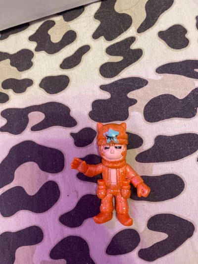 A small, orange, glitter-covered toy figurine of a character wearing a helmet with cat-like ears stands on a camouflage-patterned surface. The figurine has a blue star emblem on its helmet and appears to be a male character with a stern expression. The camouflage pattern consists of irregular black and light beige shapes. The lighting in the image has a purple hue, particularly visible on the lower left side. There is no information about the location in Vaganovo, Russia, provided in the image.