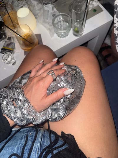 A close-up, overhead shot shows a person's legs and hand resting on a table, likely in Naousa, Greece. The table is cluttered with used drinks and what appears to be cigarette packs, suggesting a bar or restaurant setting. The person's hand, adorned with white acrylic nails and a silver ring, is holding a shimmering silver bag. The background reveals more glasses and a glowing lamp, adding to the ambiance of a night out.
