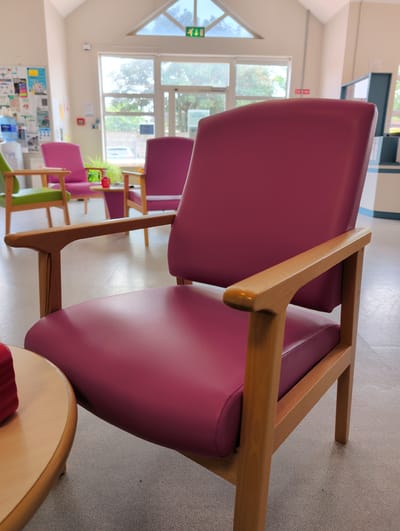 The image shows a row of empty waiting room chairs. The chairs are upholstered in a deep magenta color and have wooden frames. Some chairs are fully visible, while others are partially obscured. In the background, there are large windows that offer a view of trees and greenery, suggesting a daytime setting. Natural light filters into the room, illuminating the floor and the chairs. The room appears to be a waiting area, possibly in a hospital, clinic, or care facility, given the presence of informational posters on a wall and what appear to be medical supplies on a counter in the distance. There are no people visible in the image. The overall impression is one of quietness and anticipation, common in waiting areas. The location is identified as Waterford, Ireland.