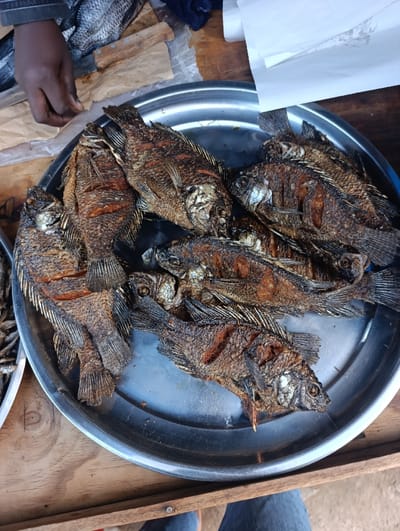 A silver platter holds multiple whole fried fish, arranged overlapping each other. The fish are golden-brown with visible scales and fins. To the left, a hand hovers over a nearby container of smaller fish. A sheet of white paper rests on the upper right. The platter sits on a wooden surface.