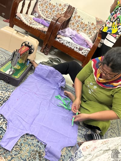 The image depicts a woman seated on a patterned rug in what appears to be a domestic setting. The woman, wearing a green top and a colorful scarf, is focused on measuring a light purple garment with a green measuring tape. A sewing machine is positioned nearby on the floor. To the left of the woman is a chair with floral upholstery, partly covered with folded clothing. Another person is partially visible in the upper left corner, wearing a shirt with cartoon characters. The location is believed to be in the city of Zirakpur, India.