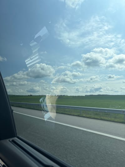 The image depicts a view from inside a vehicle, likely a car, on a highway. The foreground shows part of the car's interior, specifically the window frame and a portion of the dashboard. The highway is visible through the window, with a grey asphalt road surface marked by a white lane divider. Adjacent to the road, there is a metal guardrail and a strip of green vegetation, possibly grass or farmland. The sky occupies a significant portion of the frame and is filled with white, puffy cumulus clouds against a blue backdrop. A reflection of the vehicle's interior is visible on the window, obscuring some of the sky. The location cannot be identified without additional information. The city and country of Malenovo, Bulgaria, cannot be verified from the context of the provided image.