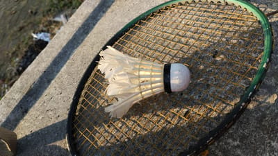 In Trosobo, Indonesia, a worn badminton racket with a green frame and yellow strings rests on a rough, grey concrete surface. A single, white shuttlecock, showing signs of wear with some feathers slightly bent, is positioned on the strings of the racket. The scene is illuminated by sunlight, casting long shadows from the racket and the shuttlecock across the concrete. The background consists of a grassy area with some debris, suggesting an outdoor setting.