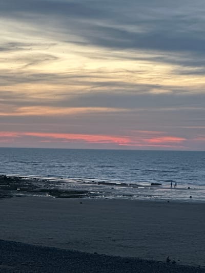 The image depicts a serene sunset scene at the beach in Veules-les-Roses, France. The sky is painted with soft hues of orange, pink, and grey, transitioning into a darker blue towards the top. Wispy clouds stretch across the horizon, creating a dynamic texture. The sea is calm, with gentle waves lapping at the shore. In the foreground, a sandy beach transitions into a rocky shoreline. A few silhouetted figures are visible near the water's edge, appearing to be a small group of people walking along the beach. The overall atmosphere is peaceful and tranquil, characteristic of a coastal sunset.