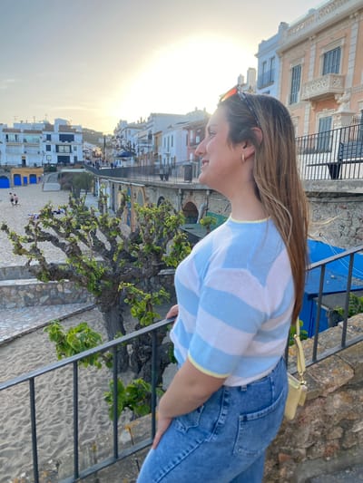A photograph shows a woman in a striped blue and white shirt and jeans leaning over a black iron railing, looking down towards a beach and town. She has long brown hair and is wearing sunglasses on her head. The beach has light-colored sand, and several people are visible on it. Blue beach umbrellas are also present. The town is composed of white buildings with a mix of architectural styles. The photo appears to be taken during the late afternoon, as the sun is low in the sky, creating a bright glow. In the foreground, between the woman and the beach, is a tree with green leaves. A stone wall and steps lead down towards the beach. In the upper background, a building with ornate architectural details is visible. The photograph's location is Calella De Palafrugell, Spain.