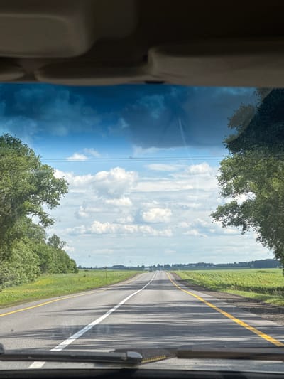 A point-of-view shot from inside a vehicle shows a highway stretching into the distance under a partially cloudy sky. The road is paved with asphalt and features a white center line and a yellow line on the right edge. Lush green trees line both sides of the road, with fields of crops visible beyond them. The sky is a mix of blue and white clouds, with some darker clouds suggesting a potential for rain. The scene appears to be set in a rural area, likely within the city of Oktyabrsky Sel'skiy Sovet, Belarus, given the landscape and road infrastructure. There are no people or animals visible in the image.
