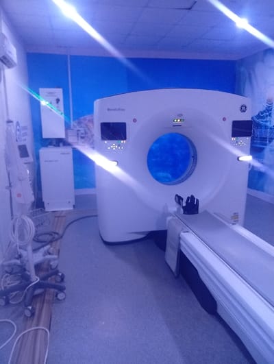 This image depicts the interior of a medical imaging room, specifically located in Okengwe - Obehira, Nigeria. The central focus is a large, white GE Revolution brand CT scanner, featuring a circular gantry opening illuminated with a blue light. The scanner's front panels display various illuminated buttons and digital readouts. A patient table extends horizontally from the base of the scanner.
To the left, a mobile medical equipment cart with multiple wheels is positioned on the floor, holding coiled white cables and other ancillary devices. Further left, an air conditioning unit is mounted high on the wall. Below it, a tall, white medical console or cabinet is visible, with a label partially reading "NATIONAL HOSPITAL ABUJA," alongside a smaller, rectangular white unit underneath. The walls of the room are painted a solid blue in their upper sections. The right wall features a large, light-toned mural or graphic depicting abstract patterns, possibly related to water or technology, with some indiscernible text. The floor is covered with a light gray or blue-gray material, with a strip of wood-patterned flooring visible along the far left wall. The room is illuminated by overhead lights, casting a cool, blue tint over the scene and producing significant lens flares across the image. No individuals are present in the room.