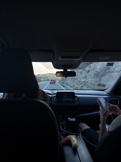 The image depicts the interior of a vehicle, viewed from a rear passenger seat, looking forward. The scene is set on a two-lane asphalt road in or around La Misión, Baja California, Mexico, characterized by hilly terrain.

In the foreground, a partially peeled banana with a yellow peel and brown blemishes rests on the dark center console armrest. A white charging cable is visible, connecting to a light-colored smartphone held by the front passenger. This passenger, whose arms and lap are visible, wears dark shorts and actively uses the smartphone with both hands. In the driver's seat, a person with long, dark hair is seen from behind, with their hands on the steering wheel. Their eyes and forehead are reflected in the rearview mirror.

The vehicle's dashboard is dark, featuring a central infotainment screen displaying "RADIO." Below the screen, a smartphone is cradled, and a silver insulated cup occupies a cup holder. The windshield ahead exhibits multiple cracks, primarily arcing across the passenger side. Through the windshield, a red SUV is visible in the distance, traveling in the same lane. To the right of the road, a steep, rocky hillside rises. To the left, a more distant landscape of green hills and vegetation is visible under a bright, possibly early morning or late afternoon sky. A blue road sign with a white symbol is partially obscured on the left shoulder.