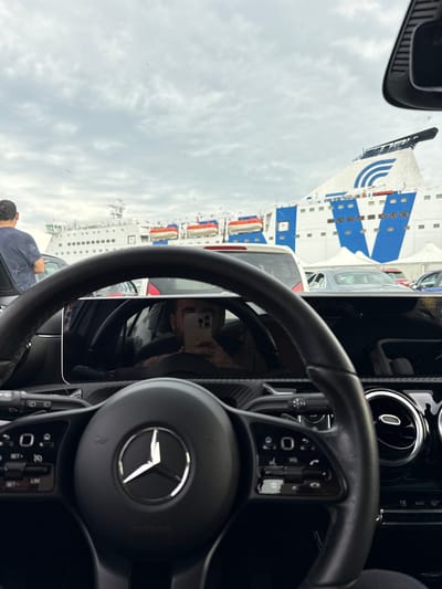 The image, taken from inside a Mercedes-Benz car in Civitavecchia, Italy, shows a ferry ship docked at the port. The steering wheel of the car is prominently displayed, with the Mercedes logo visible. A reflection in the car's infotainment screen shows a person holding a smartphone, presumably taking the photo. Outside the car, other vehicles are visible, suggesting a line of cars waiting to board the ferry. A man dressed in a navy-colored shirt is seen near the left front of the car. The sky is overcast, suggesting cloudy weather conditions. The large white and blue ferry is the dominant feature in the background.