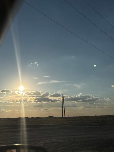 The image captures a rural landscape under a vast, partly cloudy sky at sunset. In the foreground, a dirt or gravel road stretches into the distance, with the hood of a vehicle visible at the bottom, suggesting the photo was taken from inside a car. The horizon is marked by a low, undulating terrain covered in dry grass and scattered vegetation. Several power poles and transmission lines are visible, stretching across the scene and disappearing into the distance. The sun is low in the sky, casting a warm, golden light and creating a lens flare that streaks vertically down the image. There is a bright, circular object with a greenish tint in the upper right quadrant of the sky, likely a lens artifact or a reflection. The overall impression is one of a remote, open space during the golden hour. The location is specified as Töretam, Kazakhstan.