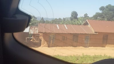 An eye-level shot from inside a vehicle shows several simple, earthen houses with corrugated metal roofs along a dirt road. Trees fill the background, with hazy mountains visible further in the distance. The houses appear to be made of mud or clay. The vegetation is sparse near the road. The shot has a slightly obstructed view due to the vehicle's window.