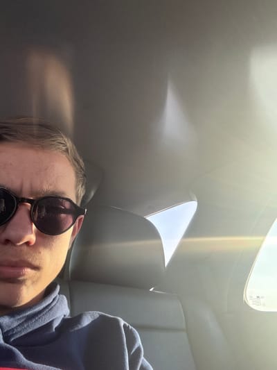 A male individual is depicted occupying the left portion of a passenger vehicle's interior in Georgi-Dobrevo, Bulgaria. He is positioned in what appears to be a front seat, wearing a dark blue hooded sweatshirt and dark-lensed, round-framed sunglasses. His facial expression is neutral. The car's interior features light-colored upholstery on the seats and ceiling. Strong, direct sunlight enters from the right side of the vehicle, creating significant lens flare across the image and brightly illuminating the interior surfaces, including portions of the ceiling and the visible rear passenger seat headrest. The sky visible through the windows on the right side appears clear and bright.