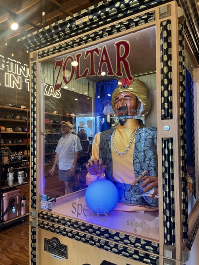 A vintage Zoltar fortune teller machine stands inside a shop, featuring a turbaned animatronic figure with a glowing blue orb in its hand. The machine reads "Zoltar Speaks $1.00," encased in a wooden frame with decorative patterns. Reflections show a man wearing a hat and shelves filled with hats and merchandise in the background. The machine appears to be in a retail setting.
