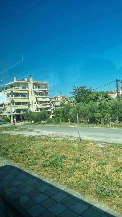 The image depicts a street scene in Larissa, Greece, under a clear blue sky. In the background, a multi-story apartment building with beige awnings and balconies dominates the left side of the frame. Adjacent to it, other buildings with similar architectural styles are visible. A road runs horizontally across the middle ground, with a grassy verge and trees bordering it. The verge on the left side of the road is lined with olive trees and other foliage. A "STOP" sign is visible on a pole near the sidewalk on the left. The foreground shows a tiled walkway, casting a shadow across a patch of dry grass. The overall impression is of a residential area on a sunny day.