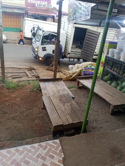 The image captures a vibrant roadside scene in what appears to be a market or commercial area in Kangema, Kenya, likely during the daytime under overcast skies.

In the foreground, two rustic wooden benches or display platforms are positioned on a dirt ground, suggesting makeshift market stalls. A bright green pole stands upright between them. To the far right, a market display rack holds pineapples, and a pile of green watermelons is visible below. The very bottom left corner shows a fragment of a tiled floor with a herringbone pattern.

The midground is dominated by a white box truck, parked diagonally on the side of a paved road. Its rear cargo doors are open, revealing several sacks of goods inside, indicating an unloading activity. A person wearing a blue shirt is visible near the rear of the truck, seemingly involved in the delivery. The truck has a "smart" logo on its front side. A series of wooden planks forms a temporary walkway or ramp leading from the street level towards the market stalls.

In the background, behind the truck, a person in an orange shirt walks on the sidewalk. Several shopfronts line the street. Prominently displayed on one building is the word "EBENEZER," repeated multiple times. Another sign reads "NELSAM SUPERMARKET." Most of the shop entrances appear to be secured with metal roll-down doors. The overall scene depicts a busy local commercial area with goods being delivered and market operations underway.