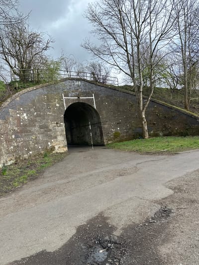The image shows a curved brick structure with a dark, arched tunnel opening next to a paved path. The structure has white painted lines forming a rectangle around the arch's top portion. Above the structure is bare trees. The path is light grey and is located on the right. The scene appears to be in a natural environment, possibly near a canal or waterway in Dunfermline, United Kingdom. The image does not contain any people.