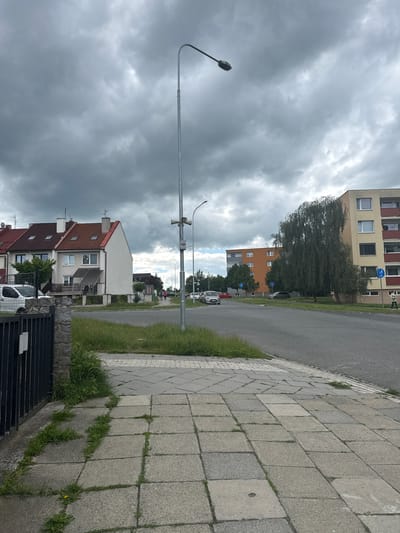 The image depicts a street scene in Olomouc, Czechia, under a cloudy sky. In the foreground, a paved sidewalk with grass growing between the stones leads to a black metal fence. Beyond the fence are several terraced houses with red tile roofs and dormer windows. A white car is parked in front of one of the houses. Further down the street, a light pole with two speakers is visible. Across the street are two apartment buildings, one orange and one yellow and beige with red balconies. Trees line the street, and several cars are parked and driving. A person is walking on the sidewalk near the apartment buildings, and a bicycle path sign is visible. The overall atmosphere is overcast and urban.
