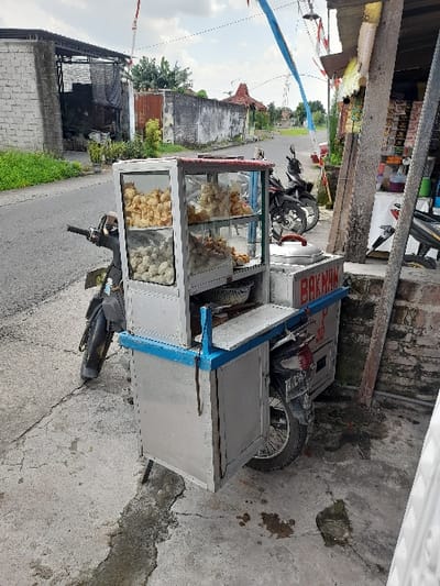 The image shows a food cart parked on a paved street in Kecamatan Purwoasri, Indonesia (latitude: -7.635, longitude: 112.136). The cart is a modified motorcycle with a metal frame and glass shelves, displaying a variety of fried food items. The cart is parked facing the street, with the front wheel of the motorcycle visible. The street is narrow and lined with brick buildings on both sides. The cart is labeled "Bakwan," a type of Indonesian fried snack. The scene is quiet, with no people present. The weather appears overcast, and the ground is dry and dusty. There is an open-air structure behind the cart, potentially a shop or dwelling, with various items visible on its walls.  There is also a partially obscured motorcycle parked nearby.  The scene is typical of a street in Indonesia, with a small business operating on a residential street.