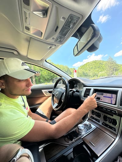 The image shows a young man driving a car, likely a Jaguar, through a scenic, hilly area with lush green trees. He is wearing a white Adidas baseball cap and a neon yellow t-shirt. His right hand is touching the car's touchscreen navigation system, which displays a map. The interior of the car features wood-grain trim and a modern dashboard. The exterior environment suggests a sunny day with a clear blue sky and some white clouds. While the specific location is not visible in the image, the context of Crkvine, Montenegro, implies a mountainous or coastal region with significant natural beauty, which is consistent with the backdrop.