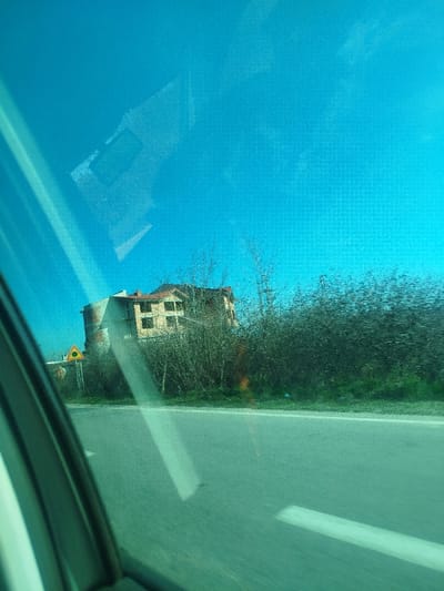 The image depicts an exterior scene in Gorno Aleksandrovo, Bulgaria, observed through a tinted vehicle window. The upper half of the frame is dominated by a clear, bright turquoise-blue sky. The lower foreground shows a grey asphalt road with a faint white lane marking. Prominent reflections of the vehicle's interior, including linear and rectangular shapes, are visible across the tinted window surface. On the left, a portion of the vehicle's dark-colored door or window frame is present.
Adjacent to the road, a yellow triangular warning sign with a black border and an indistinct circular black symbol is mounted on a pole. Behind this sign and extending horizontally is a dense growth of bare deciduous vegetation, consisting of leafless bushes and trees. Elevated behind this vegetation, a multi-story building with a light-colored facade, possibly brick or stucco, is visible. It features multiple dark, rectangular window openings and a section of a red-tiled roof. A distinctive architectural element is a curved or sharply angled corner section on the building's left side. No individuals are visible in the scene. The lighting indicates daytime conditions.