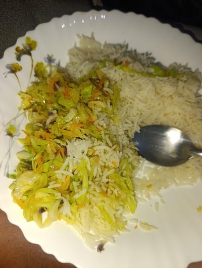 A white plate holds a serving of cooked white rice alongside a vibrant mix of sautéed cabbage and shredded carrots. The cabbage mixture appears slightly oily and yellowish-green. A stainless steel spoon rests on the rice. Floral decorations adorn the side of the plate. The food sits on a dark surface.