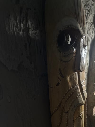 The image is a close-up, low-angle shot of a wooden mask with a weathered appearance, positioned in what appears to be a dimly lit, textured environment. The mask is predominantly brown and cream-colored, with carved features including a prominent nose, two large circular eye sockets, and what seems to be a stylized mouth area. There are decorative circular indentations or painted dots along the cheek and neck area of the mask. Fine strands of cobweb are visible, particularly within the left eye socket and stretching across parts of the mask. The background to the left of the mask is dark and appears to be a rough, textured surface, possibly a stone or concrete wall, with some areas illuminated by light creating a mottled effect. To the right of the mask, the background is lighter and also textured, suggesting a continuation of the same environment. The overall impression is one of age and perhaps neglect, with the mask being the central and sole focus of the composition. The image does not contain any people or explicit actions.

The location in Slov'yanoserbs'k, Ukraine, is not discernible from the visual elements within the image itself.