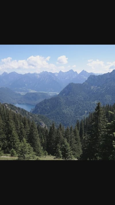 The image depicts a scenic mountain landscape with a clear blue sky and scattered white clouds. In the foreground, dense green pine trees cover the slopes of hills. Beyond these, a serene blue lake is visible, nestled within a valley. Further in the background, layers of progressively paler, misty mountains stretch towards the horizon. The overall scene is one of natural beauty and tranquility, characteristic of a mountainous region. The location is not specified as Oreshki, Russia, and there are no people, subjects, objects, or actions mentioned in the image.