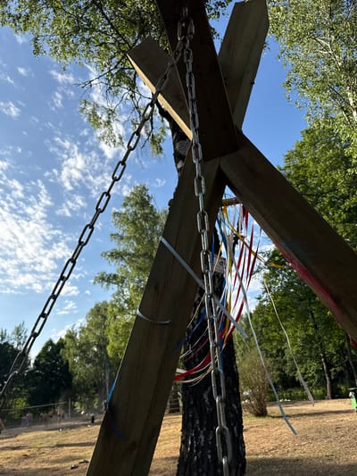 The image captures a low-angle view of a wooden structure with chains, adorned with colorful ribbons, set against a backdrop of a bright blue sky with scattered clouds and lush green trees. In the distance, a sandy area is visible with several people engaged in what appears to be a game of volleyball. The ground is covered in dry grass and some scattered debris. The overall atmosphere suggests an outdoor recreational area, possibly a park or campsite, within the Глівінскі сельскі Савет, Belarus. The focus is on the overhead structure, with the activities of people in the background serving as context for the location.