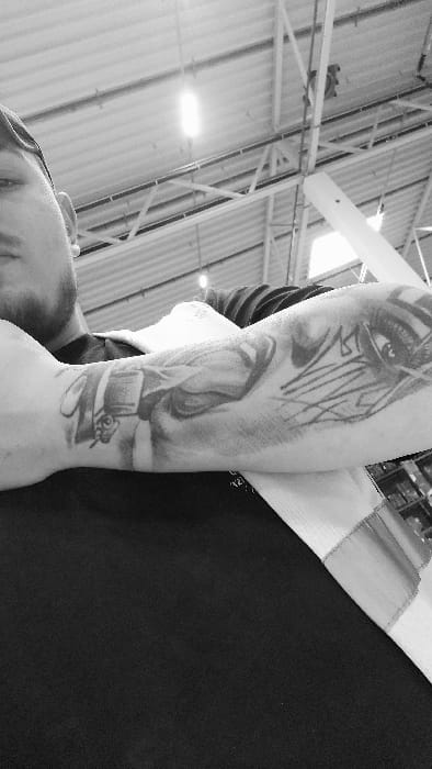The monochrome image captures a low-angle perspective inside a large commercial or industrial building in Senec, Slovakia. The primary subject is the left forearm of an individual, occupying the lower half and left side of the frame, adorned with extensive tattoos. The forearm displays a detailed portrait-style tattoo featuring a figure with distinct facial features, and further up the arm, an abstract design possibly incorporating stylized letters or an eye. The individual is wearing a dark, short-sleeved garment. A partial view of the individual's face, including a beard, jawline, and ear, is visible in the upper-left corner. The background features a high, corrugated metal ceiling with exposed structural steel trusses and beams. Multiple linear fluorescent light fixtures are suspended from the ceiling, with one prominent, brightly illuminated fixture visible in the upper-middle. A thick, white, angled support beam or column is visible on the right side of the background, extending diagonally. The environment suggests a large retail outlet or warehouse.