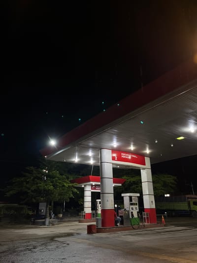 A nighttime photograph captures a Pertamina gas station in Seunebobaru, Indonesia. The scene is illuminated by the overhead lights of the gas station canopy and the ambient darkness of the night. The ground is wet, indicating recent rain or a recent washing of the area.
In the foreground, a gas pump is visible with its dispensing nozzles. A person, dressed in a red shirt and blue jeans, is seated at the pump. To the right of the pump, a green truck with the Pertamina logo is parked.
The gas station canopy is red and white, with several lights illuminating the area. The structure is supported by white pillars. Signs on the canopy indicate "PERTAMAX TURBO" and "PERTAMAX". A large green truck is parked further back on the right side of the frame.
Trees are visible behind the gas station. The sky above is dark and appears to have some rain streaks, visible as faint vertical lines. The overall atmosphere is one of a typical, operational gas station at night.