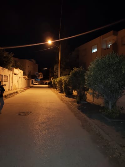A dimly lit street scene at night in an unknown location in Tunisia. A street lamp illuminates a paved road that curves into the distance. On the left, a white wall with barred windows borders the street, and a person in dark clothing and light pants is partially visible on the left edge of the frame. Further down the street, a white car is parked. On the right side of the street, a row of lush green bushes and trees lines the pavement. Buildings with illuminated windows are visible in the background, suggesting residential properties. The sky is dark, indicating nighttime, and a thin beam of light from the street lamp stretches across the sky. The road surface appears wet or reflective in places, possibly due to recent rain or dew, and reflects the orange glow of the street light. A circular manhole cover is visible on the road in the foreground.
