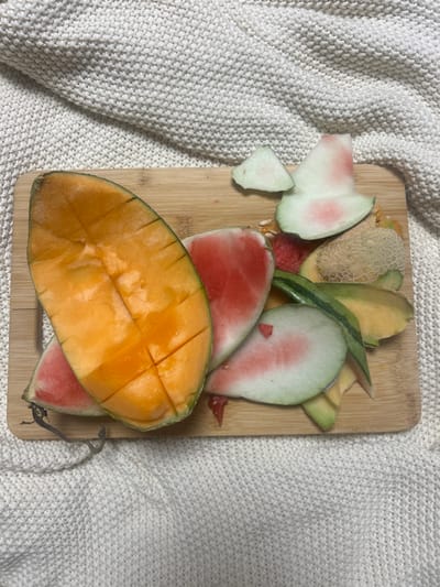 The scene depicts a rectangular wooden cutting board, approximately 30-40 cm in length, positioned diagonally on a light-colored, possibly cream or off-white, knitted textile surface. The cutting board contains remnants of consumed fruit. Prominently featured on the left is a large, segmented half of an orange-fleshed cantaloupe (rockmelon), cut into cubes but still attached to its light green/orange rind. To the right of the cantaloupe are various pieces of watermelon rind, predominantly white with remnants of pink flesh and some green outer rind, interspersed with several dark watermelon seeds and small red fragments of watermelon flesh. Also present are pieces of pale tan/orange cantaloupe rind, exhibiting its characteristic netted pattern. The items are scattered across the board, indicating recent fruit consumption. This scene is located in Corrimal, Australia. No individuals are present, and no active interactions are occurring. The environment suggests an indoor setting.