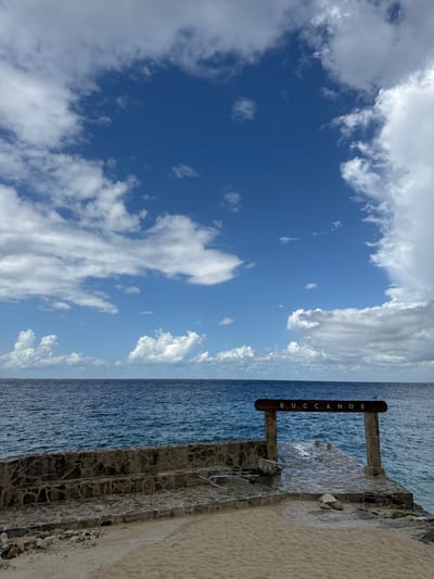 The image depicts a coastal scene in Cozumel, Mexico, specifically at Buccanos Beach Club and Grill, indicated by a prominent sign. The sky occupies the upper two-thirds of the frame, showing a clear blue expanse with scattered white cumulus clouds. Below the horizon, a deep blue ocean with visible surface ripples extends across the middle ground. In the foreground, from bottom-left to bottom-right, a light-colored sandy area transitions into a stone-built structure. This structure consists of a multi-tiered stone wall on the left, leading to a flat stone pier or jetty extending into the ocean on the right. At the entrance to this pier, a rustic wooden sign stands, supported by two vertical posts, bearing the word "BUCCANOS" in light-colored letters on a dark wood plank. A metallic, triangular-shaped object, possibly a ladder section, lies on its side on the stone steps to the left of the pier. The scene is devoid of people or active interactions.