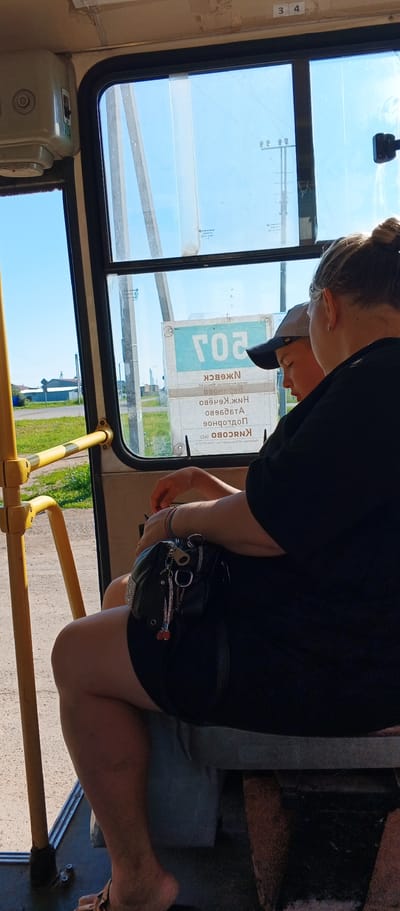 The image captures an interior view of a bus, likely in the city of Kiasovo, Russia, judging by the signage visible through the window. The focus is on two individuals seated on a bench. On the right, an adult, possibly female, wearing a black top and dark shorts, is seen from the back and side. Her arm rests on a black purse that is adorned with several charms. To her left and slightly in front, a young person, likely a boy, wearing a baseball cap and a t-shirt, is looking downwards. The bus interior features a yellow handrail on the left and a window offering a view of a sunny outdoor scene with a blue sky and some vegetation. A sign is visible through the window with text that appears to be in Cyrillic script, partially obscured. The lighting suggests it is daytime, with sunlight filtering into the bus.