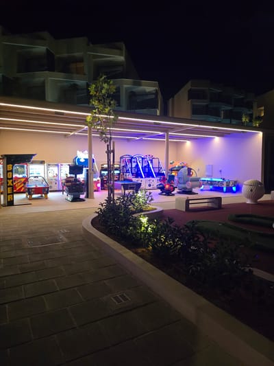 The image depicts an outdoor arcade and miniature golf area at night, located in Polis Chrysochous, Cyprus. The arcade features various game machines, including air hockey and video games, illuminated by neon lights. In the background, there is a modern building with a distinctive architectural style, possibly a hotel or resort. The miniature golf course is visible in the foreground, with green turf and golf equipment. The scene is well-lit, with a combination of artificial lights and the natural darkness of night. There are no people visible in the image.