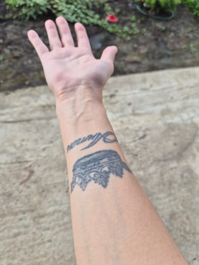 The media file shows a close-up view of a person's forearm, which has a tattoo of a crown and some cursive text. The tattoo is on the inner side of the forearm and appears to be in black ink. The text is difficult to read but seems to be a name. The skin has a light to medium tone.
The arm is positioned with the palm facing upwards and slightly to the left. The fingers are spread open, suggesting an open hand gesture. In the background, out of focus, is a patch of green vegetation and a concrete-like surface. The lighting suggests it could be daytime, possibly outdoors, given the natural light and the presence of greenery. The concrete surface has a mottled appearance with some darker and lighter patches.
The overall impression is a still shot focused on the tattoo and the hand. There are no clear indicators of a specific event or strong emotions being conveyed. The location context provided is Lenina, Russia, but there are no visual cues within the image that specifically identify this location.