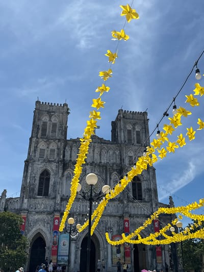 This image shows a view of the Phat Diem Cathedral in the city of Phat Diem, Vietnam. The cathedral is a large, stone building with two prominent towers. It is decorated with strings of yellow, star-shaped paper lanterns and string lights, which hang in front of the structure. The sky is blue with scattered clouds. In the foreground, several people are visible, some walking and others standing. Banners with images and text are hung on the cathedral walls. The overall scene suggests a festive or celebratory atmosphere.