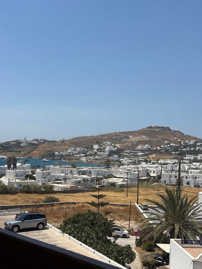 This is an outdoor, panoramic image capturing the town of Ornos, Greece, under a clear, cerulean sky. The background is dominated by a sun-baked, light-brown hill, extensively dotted with white, cubic, multi-level buildings and accessed by visible terraced roads. Immediately below the hill, Ornos Bay displays turquoise water, populated by approximately 20-30 white and light-colored vessels, including yachts and smaller boats. The shoreline and lower slopes are densely covered with numerous white, traditional Cycladic-style buildings. The mid-ground features a large expanse of dry, golden-brown scrubland. In the foreground, a light-grey SUV is parked on a paved, marked parking space on the left. A white compact car is parked on an unpaved area to the right. A prominent palm tree with dark green fronds stands on the right, alongside several smaller coniferous trees. Multiple wooden utility poles with power lines are visible throughout the townscape. The image is framed at the bottom by a dark-colored balcony railing. No human figures are identifiable.
