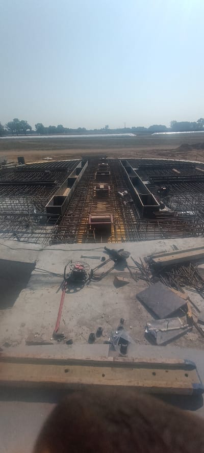 An exterior, wide-angle, eye-level shot depicts a construction site with extensive rebar framework and concrete forms laid out in a rectangular pattern, suggesting the foundation for a large structure, possibly a bridge or building. The foreground features a concrete surface with various construction tools scattered about, including a grinder, a rebar-bending tool, a spool of wire, and a plastic bag containing black objects. In the middle ground, the rebar grid is densely packed, with wooden forms creating channels and open spaces within the framework. The background shows a flat, open field with scattered trees and a hazy sky. The overall scene is one of active construction in an undeveloped or rural area. The image does not contain any specific identifiable landmarks or features that would confirm a location in Magnuszowice, Poland.