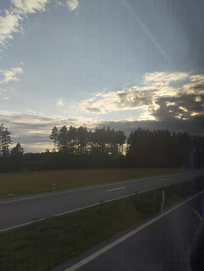 The image captures a rural scene with a highway in the foreground and a treeline in the background. The sky is partly cloudy with sunlight breaking through the clouds. The highway is asphalt with white lane markings. To the left of the highway is a grassy field, and to the right is a dark treeline. There are a few white posts along the side of the road. The image appears to be taken from a moving vehicle due to motion blur on the right side of the frame. The lighting suggests it is either early morning or late afternoon. The location is identified as Lyubonitski Sel'skiy Sovet, Belarus.