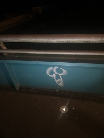 The image shows a close-up view of graffiti on a teal-colored wall at night. The graffiti appears to be a crude drawing of a penis. There are metal railings above the wall and the ground below is dark and appears to be asphalt with some small lights reflecting. The location is identified as Sempigny, France.