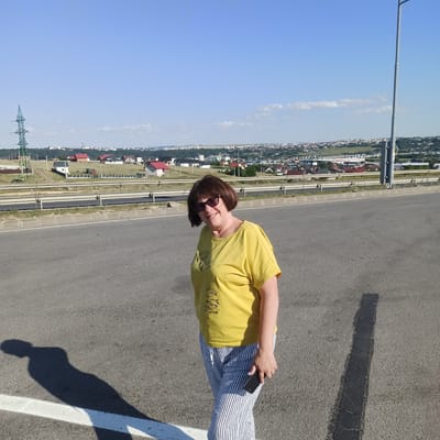 In Șcheia, Romania, a woman stands on an asphalt road, likely a highway, under a clear blue sky. She is wearing a bright yellow t-shirt with a graphic design and vertically striped pants. The woman has short, dark hair and is wearing sunglasses. She is holding a black mobile phone in her right hand and smiling at the camera. Behind her, a landscape unfolds with a row of houses and buildings stretching across rolling hills, with a prominent power line tower visible in the distance. The foreground of the image is dominated by the road surface and a shadow of the woman cast on the asphalt.