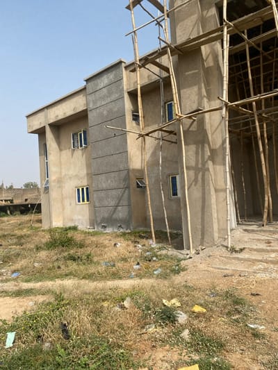 A multi-story concrete structure, approximately three levels high, is under construction in Yan Dala, Nigeria. The building's exterior primarily consists of unpainted, light-toned concrete with a rough finish. A central vertical section of the facade is covered with a darker, horizontally textured grey render. Multiple rectangular windows with blue glass panes and light-colored frames are visible, with several windows on different levels appearing partially open. Extensive, multi-level scaffolding constructed from natural wooden poles is erected along the right side and front of the building. Exposed pipes or conduits are visible on the lower part of the building's exterior. The immediate foreground consists of dry, sparse vegetation and bare earth, significantly littered with plastic bags, wrappers, and other debris. In the distant left background, the unfinished foundation or partial structure of another concrete building is visible. The sky above is clear and pale blue. No individuals, actions, or direct interactions are present in the scene.