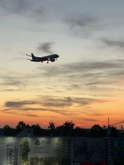 The image captures a dramatic sunset or sunrise scene in Otopeni, Romania, featuring a large airplane in flight against a vibrant sky. The sky transitions from warm orange and red hues on the right to lighter grey and white clouds on the left.

A prominent passenger jet is visible in the mid-ground, flying horizontally from left to right. It appears to be at a relatively low altitude, potentially on approach or departure. A dark exhaust plume or contrail emanates from its engines. The aircraft is silhouetted against the bright sky, with distinct red and white lights visible on its underside.

In the lower portion of the image, the ground is dark and features silhouetted trees and various urban structures. On the right side, there are large advertising banners or construction site hoardings. One significant banner displays the brand "HILS SUNRISE" along with contact information: a phone number "0310 052 174," a website "www.hils.ro," and an address "B-dul Theodor Pallady 57A." Another adjacent sign shows a graphic of a bicycle.

The scene conveys a sense of dynamic activity within an urban landscape, set against the beautiful, transient colors of dawn or dusk. A small, dark insect or bird is also visible against the cloudy sky.