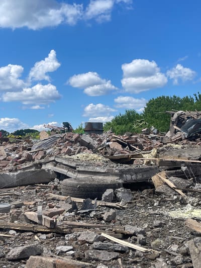 The scene shows the aftermath of a destruction event, likely an explosion, in Terny, Ukraine. The debris field consists of broken concrete slabs, shattered bricks, wooden planks, and metal fragments. There is a large, damaged tire in the foreground. The sky is blue with white puffy clouds. There are trees visible in the background, some of which appear to be damaged. The environment is largely barren and desolate. There is a sense of destruction and chaos throughout the scene. The damage appears extensive. No people are visible in the scene. There is no evidence of ongoing activity or response to the damage. The coordinates provided indicate a location in a residential area of Terny.