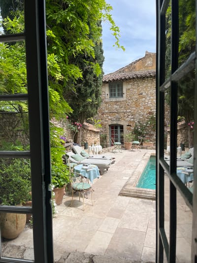 The image presents a tranquil outdoor scene, viewed through the panes of a window, looking out onto a charming courtyard.

In the midground, a traditional stone house with a tiled roof stands, showcasing rustic architecture with light-colored, irregular stones. It features a window with closed green shutters on the left side and a smaller window below it, revealing a darker interior.

To the right of the house, a rectangular swimming pool with inviting turquoise water is set into a paved patio. The patio is made of light-colored flagstones, extending across the foreground and along the pool's edge.

The courtyard is rich with lush greenery, including tall cypress trees, leafy vines, bushes, and flowering plants, some with small pink blossoms. Several terracotta pots containing plants are scattered across the patio, enhancing the garden's appeal.

Outdoor furniture is arranged on the patio, suggesting a comfortable area for relaxation and dining. This includes two round tables: one draped with a light blue tablecloth, accompanied by several chairs with light-colored, possibly striped, cushions; and another smaller table with a striped top and two chairs, one with a green cushion. An additional armchair or lounger with a striped cushion is also visible.

The scene is illuminated by bright daylight, casting shadows from the furniture, indicating a clear and sunny day, likely late morning or afternoon. The sky visible above the house is partly cloudy. No people are present in the image, and no text is visible. The overall aesthetic of the stone architecture, Mediterranean-style vegetation, and sunny ambiance aligns with the specified location of Saint-Victor-la-Coste, France, evoking a sense of peaceful, leisurely outdoor living.