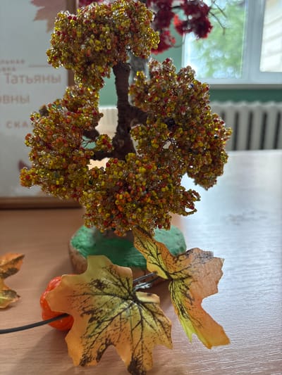 A beaded tree with colorful autumn leaves sits on a wooden table. The tree is adorned with small beads in shades of yellow, orange, red, and clear, resembling a miniature version of a tree in fall foliage. Its trunk is textured and brown, and it stands on a green, textured base. In front of the tree, there are two fabric autumn leaves, one larger and more detailed than the other. Next to the larger leaf is a small, bright orange decorative pumpkin. Behind the tree, out of focus, is a window with a view of greenery outside and what appears to be a radiator against a light blue wall. On the wall behind the tree, there is some text that is out of focus, but some letters are discernible, suggesting it might be a poster or sign. The overall setting appears to be indoors, possibly in a classroom or office, given the presence of text and the window with a radiator. The scene evokes an autumnal theme and appears to be a decorative arrangement. The location specified, Ветрынскі сельскі Савет, Belarus, is a rural administrative unit, suggesting this might be a scene from a rural school, community center, or home within that area.