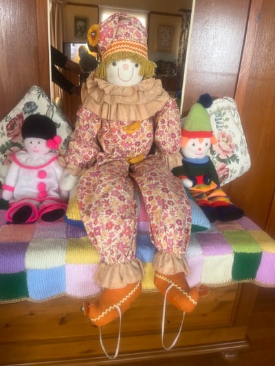 The image presents a cluttered interior scene. In the foreground, there is a handmade patchwork quilt with square sections of varying pastel colors, with gold trim. Resting on the quilt are several fabric dolls and plush figures. One is a clown doll with a floral patterned body and a ruffled collar, and another is a knitted snowman doll with a green hat. Additionally, there's a knitted doll with black hair and a pink and white outfit. These dolls are placed on floral patterned cushions or pillows.
The background features a wooden door. The location appears to be indoors, possibly a bedroom or living room.
Without additional information, it's impossible to ascertain if this scene is located in Golspie, United Kingdom.